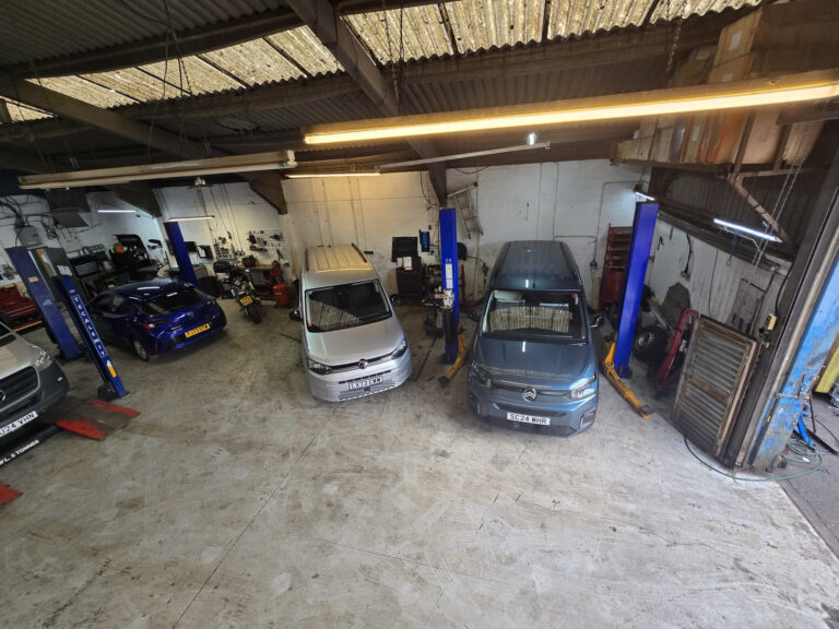 Interior view of an auto repair shop with two blue vehicle lifts and multiple cars on the floor, tools and workbenches along the walls, and overhead lighting illuminating the space.