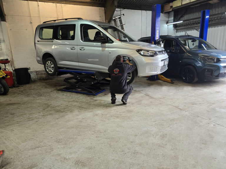 Technician kneels by the front wheel of a silver minivan on a hydraulic lift in an auto repair shop.