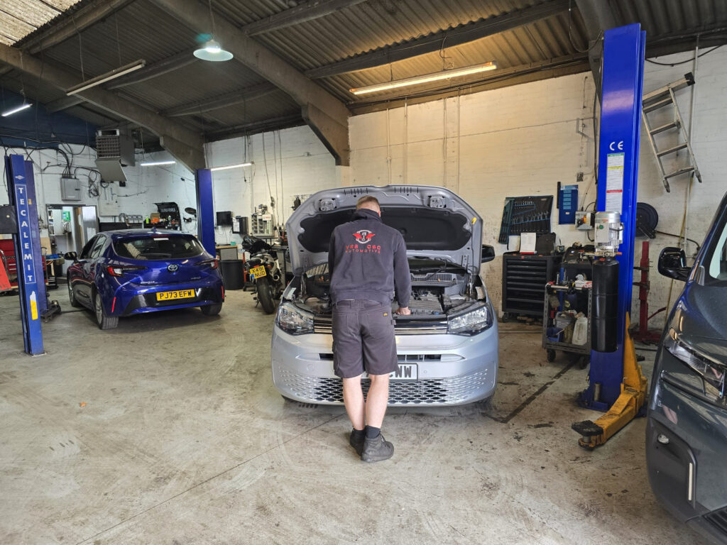 Mechanic standing at the open hood of a silver sedan in a busy auto repair shop.